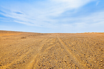 Obraz premium Barren gravel desert landscape in Dahaidao, Xinjiang, China, with tire track leading toward a low horizon beneath a blue sky, conveying remoteness and travel potential.