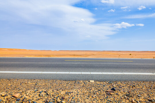 Desert highway crossing the arid Dahaidao landscape of Xinjiang, China, with gravel roadside, open steppe, blue sky, and scattered clouds.