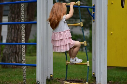 Little red-haired girl climbing a rope ladder at an outdoor playground, reaching for the next rung while wearing a pink ruffled dress, enjoying active play in a green park setting.