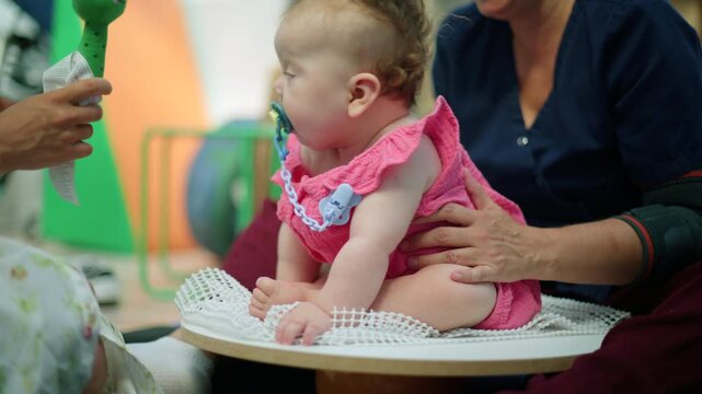 Baby girl in pink outfit on a rehabilitation ball, getting help from an adult and holding a pacifier during therapy session