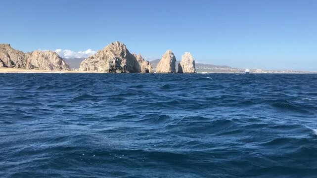 Sea level view of rolling ocean waves facing the iconic El Arco rock formation at Land's End, with rugged coastal cliffs under a clear blue sky in Cabo San Lucas, Baja California Sur, Mexico.