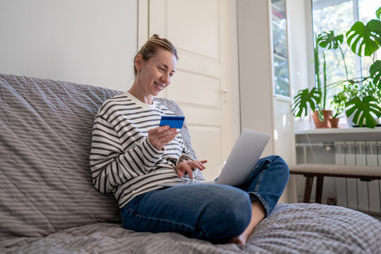 Smiling relaxed woman sitting on sofa at home using laptop and credit card for online shopping, cheerful female browsing marketplaces on computer, purchasing and making payment transactions with card.