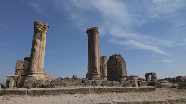 Slow ground-level moving shot between monumental Roman columns and stone walls at Volubilis, revealing an ancient passage within Morocco's UNESCO World Heritage archaeological site.