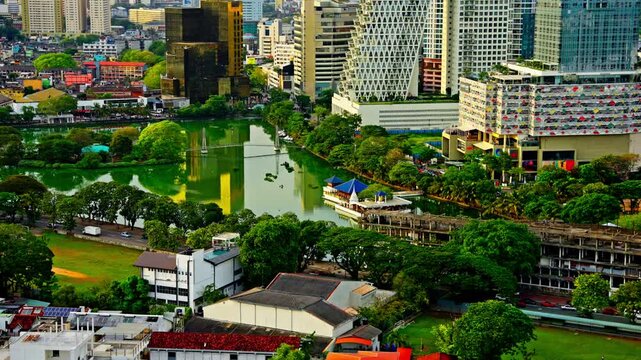 Green waters of Beira Lake mirror the surrounding hotels, offices and colorful rooftops of central Colombo, Sri Lanka, with tree-lined streets and traffic weaving along the lakeside.