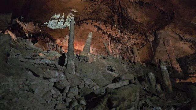 Dim limestone cave interior with stalactites, stalagmites, rocky floor, dramatic shadows, warm artificial lights, and textured mineral formations for underground nature footage.