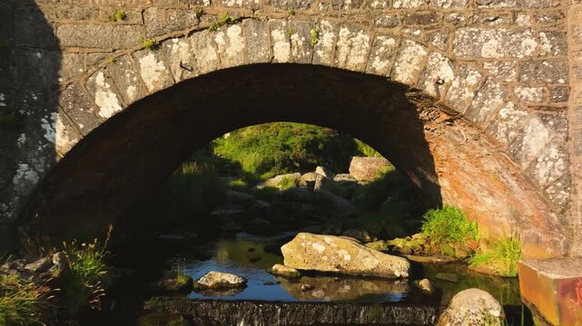 Low arch stone bridge spans a shallow rocky stream in rural Wicklow as the camera rises to reveal the sunlit hillside beyond.