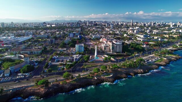 High-angle aerial drone shot pulling back and ascending over the Caribbean sea coastline, a coastal highway, the Heroes obelisk monument, and the city skyline in Santo Domingo