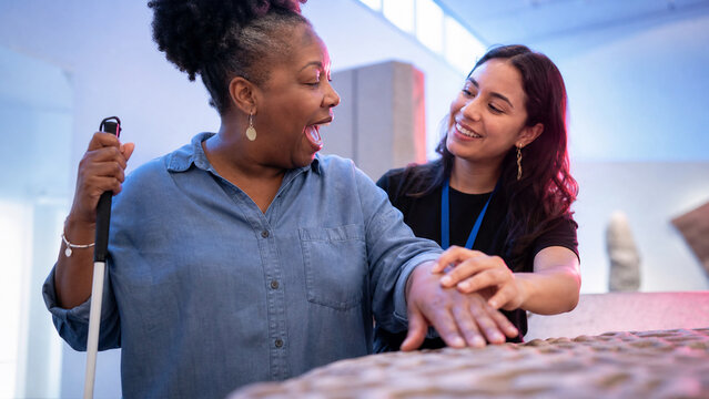 Blind woman with a guide exploring museum exhibition by touch. Smiling assistant helping visually impaired female visitor. Concept of accessibility, education, and inclusive culture for all.