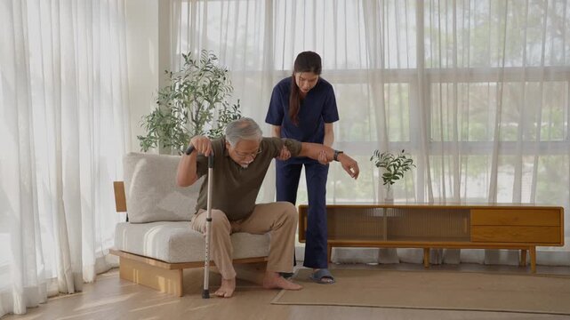 Elderly man using cane with female caregiver assistance bright modern living room, senior adult receiving support while walking and standing from chair, assisted mobility and careful movement home