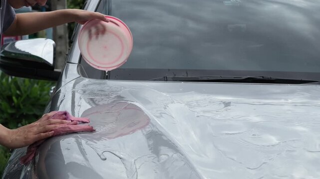 Close up of a hand pouring water from a pink basin over a soapy car hood while wiping it clean with a microfiber cloth