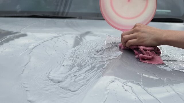 Extreme close up of a hand wiping soapy water across a wet silver car hood with a pink microfiber cloth during a hand car wash