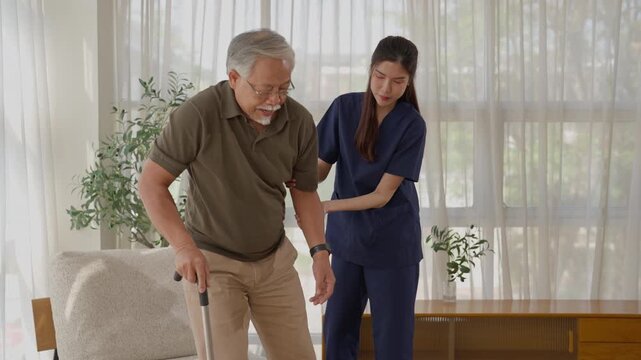 Older adult using walking cane indoors home, standing near sofa and wooden cabinet with plant, caregiver support during mobility exercise, living room mobility aid for safe senior walking assistance