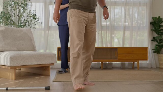 Older adult walking barefoot with caregiver bright living room, cautious movement and rehabilitation with walking cane, medical care, wooden furniture, plants, soft curtains, natural daylight
