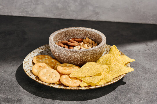 Cracker, tortilla chip and mixed nut snack platter with crisp golden textures on a dark table for party menu and recipe