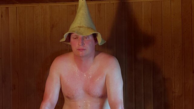 Medium close up shot showing face and chest of sweaty adult man in conical felt headwear looking exhausted while enjoying hot steam inside rustic sauna room in Baltics