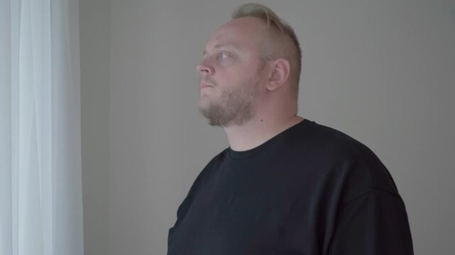 Man checks his smartphone and repeatedly looks out a bright window, suggesting anxiety while waiting for someone to arrive.