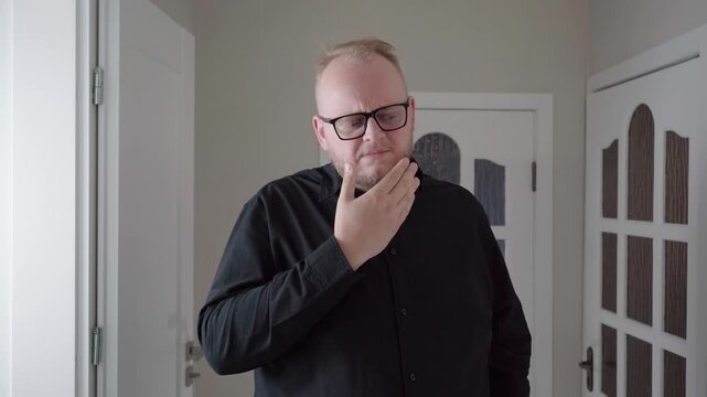 Caucasian man in glasses covers his mouth and jaw with his hand, reacting to tooth ache discomfort against a neutral indoor background.
