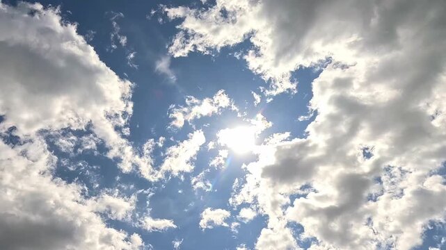 timelapse of fluffy white clouds floating across a blue sky, the bright sun shining through changing gaps as the clouds slowly merge, expand and gradually change shape throughout the frame.