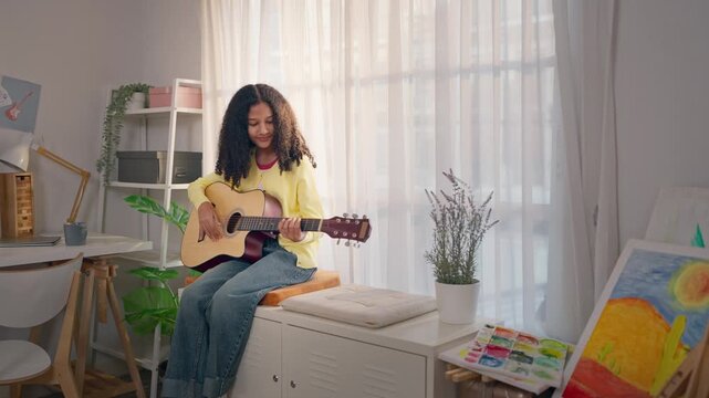 African American teenage girl plays guitar while sitting on a cabinet. 