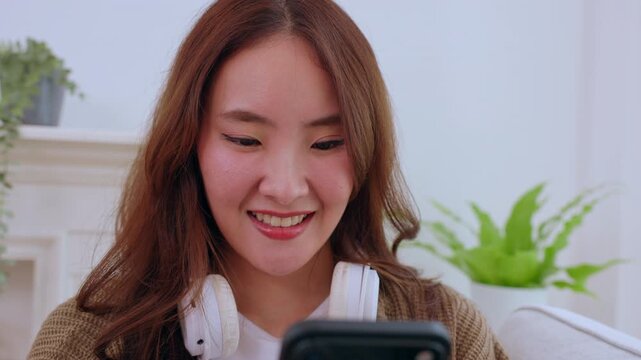 Young Asian woman smile headphone smartphone indoor home sofa plant cheerful casual technology cozy living room mobile device bright space happy adult couch