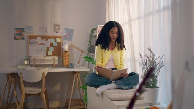 African American teenage girl sit on a white cabinet and reading a book. 