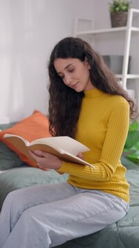 Young Latino female college student sitting on bed and reading a book. 