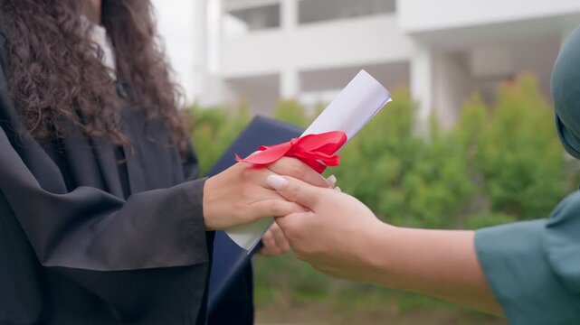 Close up of mother holding hands young daughter wearing graduation gown. 