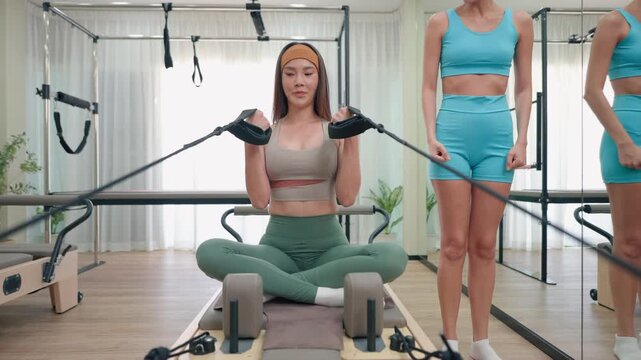 Young women exercise on Pilates reformer machine with guidance from trainer. 