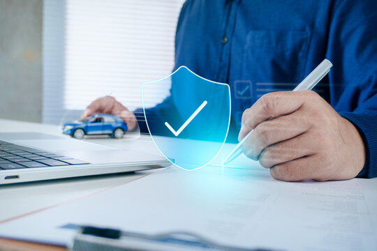 Business professional signing car insurance policy document on office desk with toy car and holographic shield checkmark icon, concept of vehicle insurance coverage, safe driving, and auto protection.