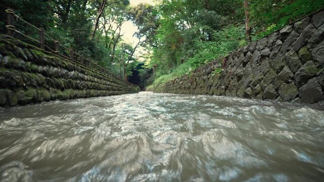 Horizontal low angle shot of a fast-flowing river between stone walls in a lush green summer forest with cicada sounds