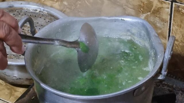 Boiling green-leafed moringa vegetable soup in a metal pot using a ladle on a gas stove.