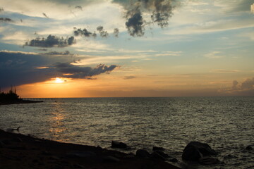 Obraz premium Tranquil seaside sunset with dark silhouettes of rocks and trees on the shore, vibrant orange and blue hues filling the sky beneath scattered clouds, reflecting on the calm ocean water.