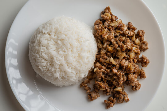 Overhead top view flat lay of Thai style stir-fried ground pork with minced garlic and pepper served with steamed rice on a round white plate.