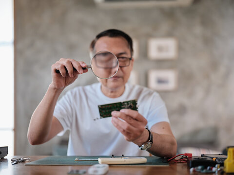 Mature Asian man looks at and considers circuit board details with magnifying glass, focusing on precision checks for home electrical appliance DIY fix, electronics component hobby, handyman workshop.