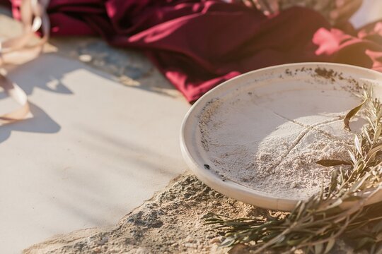 Round stone dish of ashes scored with a cross, featuring gritty textures, a deep red cloth, and olive sprigs for Ash Wednesday and parish communication