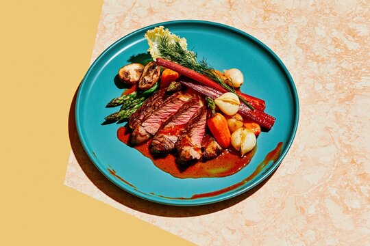 Sliced beef steak with glossy jus on a teal porcelain plate, featuring roasted vegetables and vibrant colors for restaurant menus and dining campaigns, with copy space