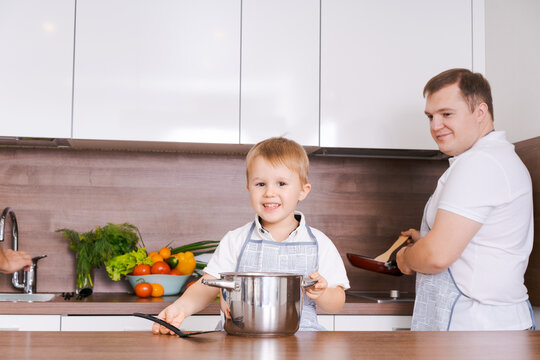 Happy caucasian boy in an apron holding a big steel pot while his father cooks with a frying pan in a white kitchen, ideal for family, home cooking and childhood content