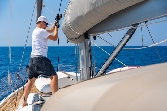 Man trimming mainsail on a sailing yacht during offshore voyage. Experienced sailor adjusting sail shape for efficient wind propulsion, safe navigation, boat control and blue water cruising lifestyle.