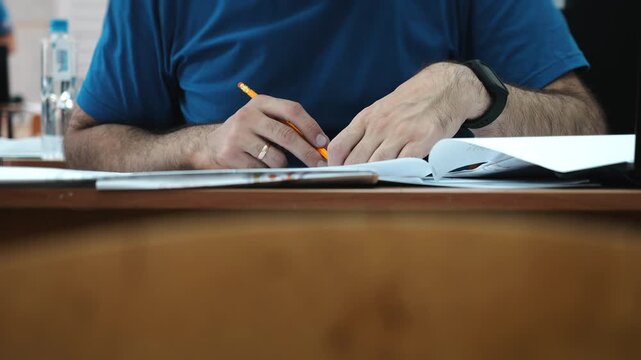 Close-up of man writing with pencil and filling out paperwork at desk. Hands working with documents, forms and records in office or professional setting.