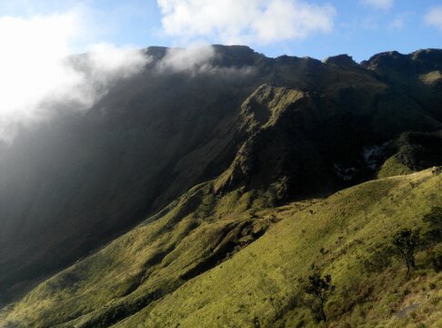 green mountain ridge with clouds rolling over volcanic slope, sunlit crater rim, majestic hiking landscape in Indonesia