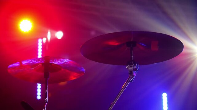 Close-up of drum cymbals on a concert stage illuminated by colorful spotlights. Live music equipment in atmospheric red and blue stage lighting.