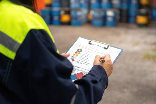 Checking on chemical hazardous safety checklist form, close-up at worker hand, with factory warehouse and metal barrel storage as background. Industrial safe work practice scene, selective focus.