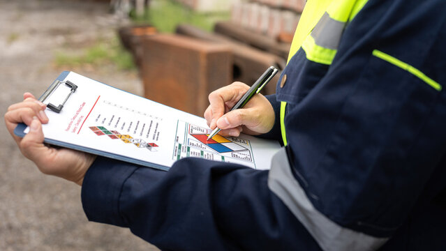 Checking on chemical hazardous safety checklist form, close-up at worker hand, with factory warehouse and metal barrel storage as background. Industrial safe work practice scene, selective focus.