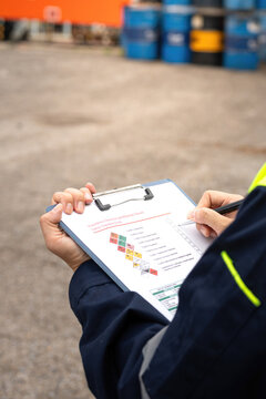 Checking on chemical hazardous safety checklist form, close-up at worker hand, with factory warehouse and metal barrel storage as background. Industrial safe work practice scene, selective focus.
