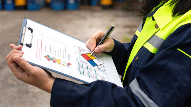 Checking on chemical hazardous safety checklist form, close-up at worker hand, with factory warehouse and metal barrel storage as background. Industrial safe work practice scene, selective focus.