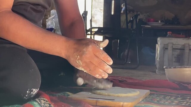 A person's hands shaping fresh dough on a wooden board, with several rounded dough portions resting on a colorful traditional rug in a rustic setting.