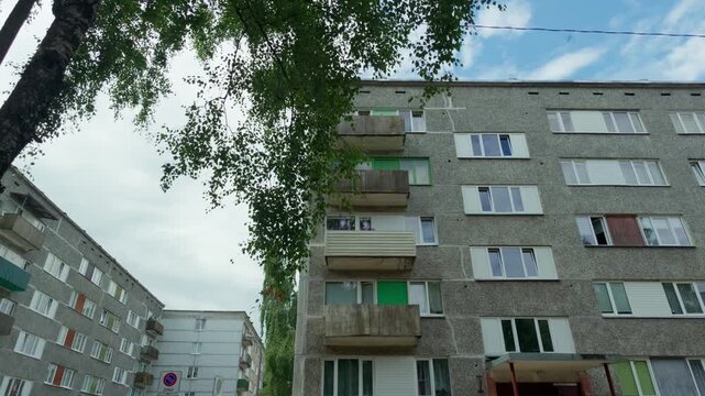 Static wide shot displaying grey concrete Soviet block apartment building with colorful balconies framed by green birch tree branch in Daugavpils, Latvia, daytime scene.