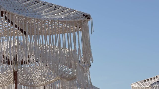 A textured macrame canopy stretches across the frame with a white building and clear sky in the distance