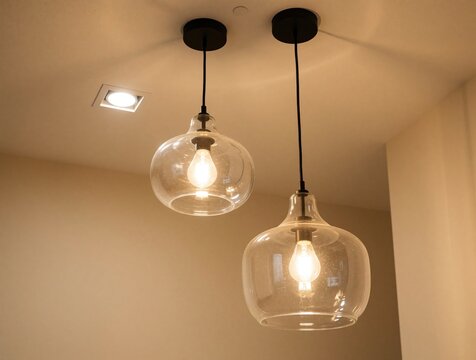 Two modern glass pendant lamps hang from white ceiling. Clear bulb light fixtures glow in warm interior room. Minimalist hanging light decor provides bright illumination in home cafe. Stylish glass