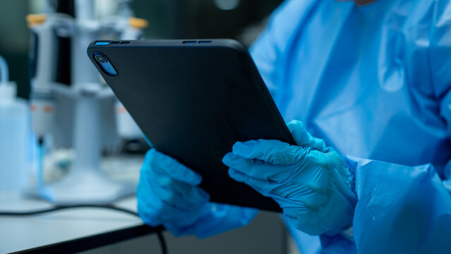 Gloved lab worker holding a tablet in a medical laboratory with blurred equipment in the background.